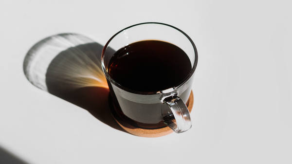 A clear glass cup filled with a dark liquid, presumably coffee, placed on a saucer. The cup and saucer are on a surface with a distinct shadow cast by the light source, creating an interesting contrast of light and shadow.